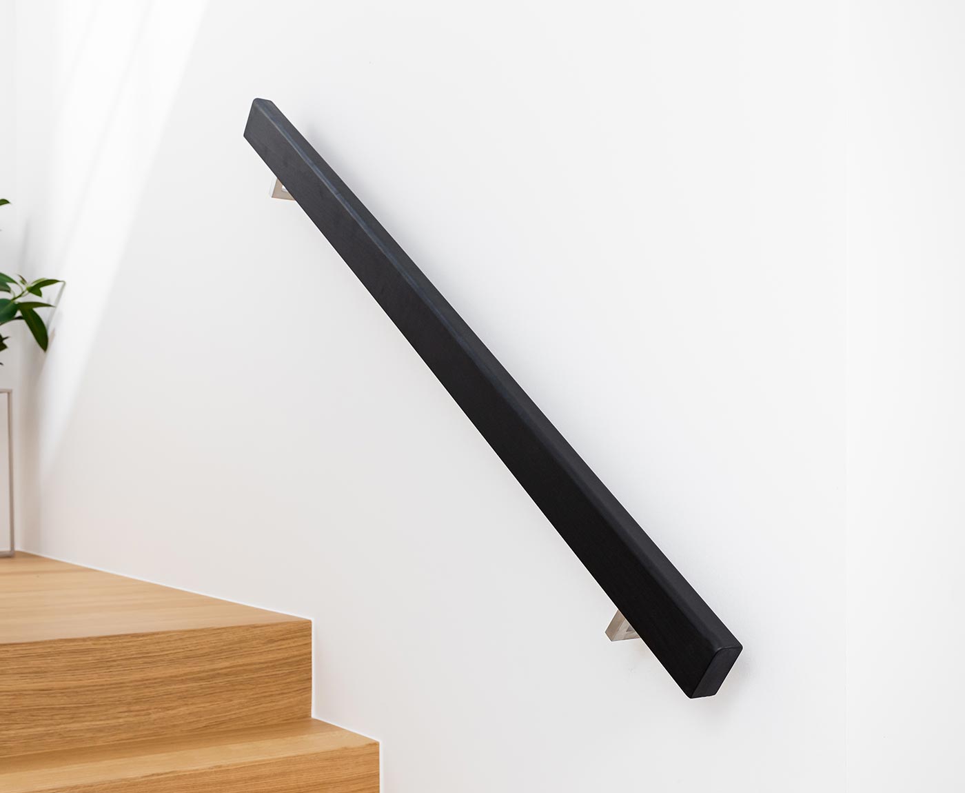 Schwarzer, rechteckiger Handlauf aus Holz an weißer Wand, mit Handlaufhaltern aus Edelstahl.